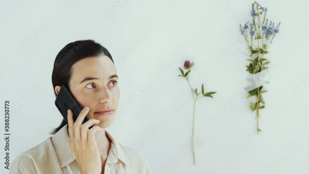 Chest up studio shot of attractive brunette woman speaking on mobile phone while standing against white wall decorated with flowers Chest up studio shot of attractive brunette woman speaking on mobile phone while standing against white wall decorated with flowers