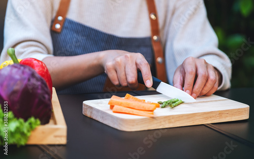 Closeup image of a woman cutting and chopping asparagus and carrot by knife on wooden board