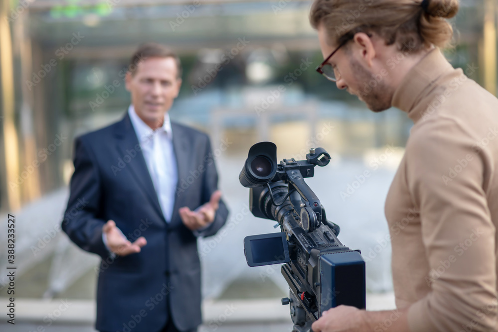 Long-haired operator filming mature male reporter in front of fountain