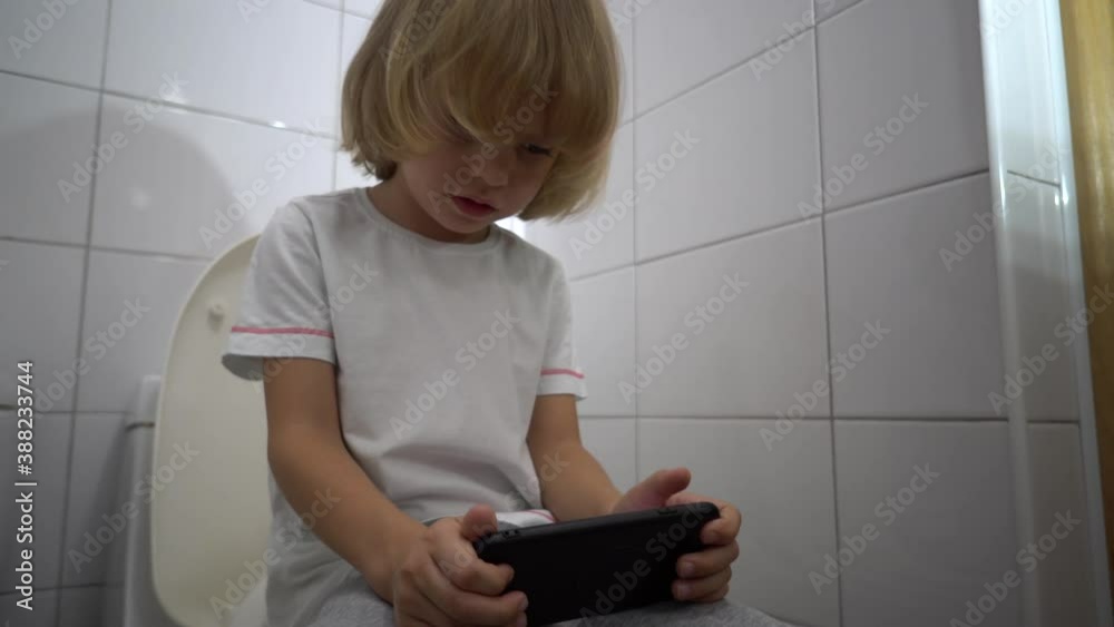 Handsome blond 5-aged boy playing game or watching cartoons on his mobile phone while sitting on the toilet bowl. Handsome blond 5-aged boy playing game or watching cartoons on his mobile phone while sitting on the toilet bowl.