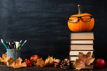 Pumpkin with glasses, halloween concept, books, pens, fallen leaves, on the table, on background of a chalkboard. Template concept of autumn mood.