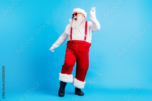 Full length photo of crazy funky santa claus with big stomach beard dance x-mas christmas discotheque wear suspenders overalls sunglass headwear isolated blue color background photo