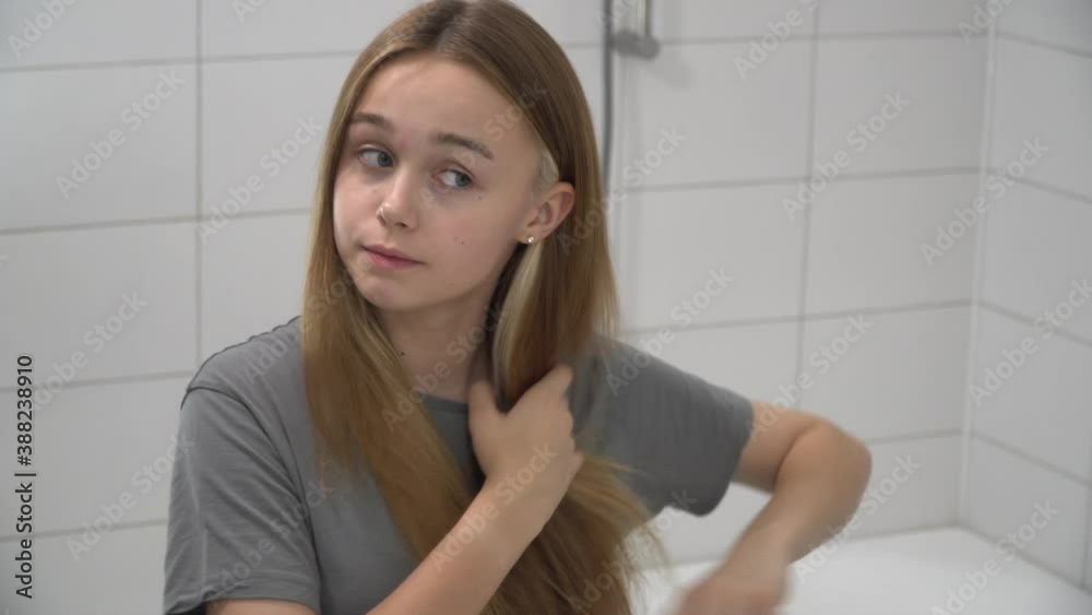 Attractive happy confident young beautiful young woman brushing healthy long hair with wooden hairbrush looking in the mirror. Teenage girl doing hairstyle in bathroom in the morning at home get ready Attractive happy confident young beautiful young woman brushing healthy long hair with wooden hairbrush looking in the mirror. Teenage girl doing hairstyle in bathroom in the morning at home get ready