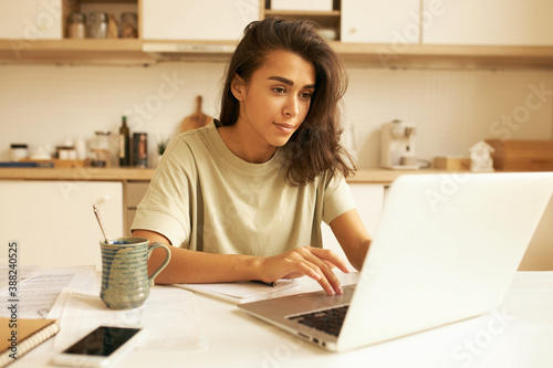 Indoor image of beautiful concentrated young female translator sitting in kitchen with mug, papers and portable computer translating technical document using free internet connection at home