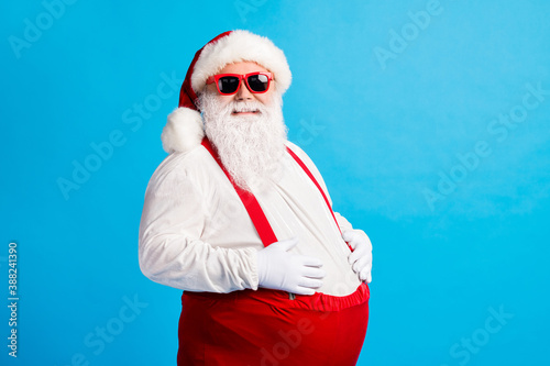 Photo of crazy overweight santa claus touch his big belly enjoy x-mas christmas newyear tradition feast wear cap suspenders overalls isolated over blue color background