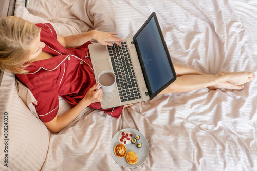 Soft cozy photo of slim tan woman in red pajamas on the bed with laptop, cup of cocoa and breakfast, top view point.