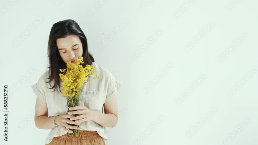 Studio waist up shot of beautiful brunette woman standing against white background, holding bouquet of yellow flowers, smelling it and looking at side pensively
