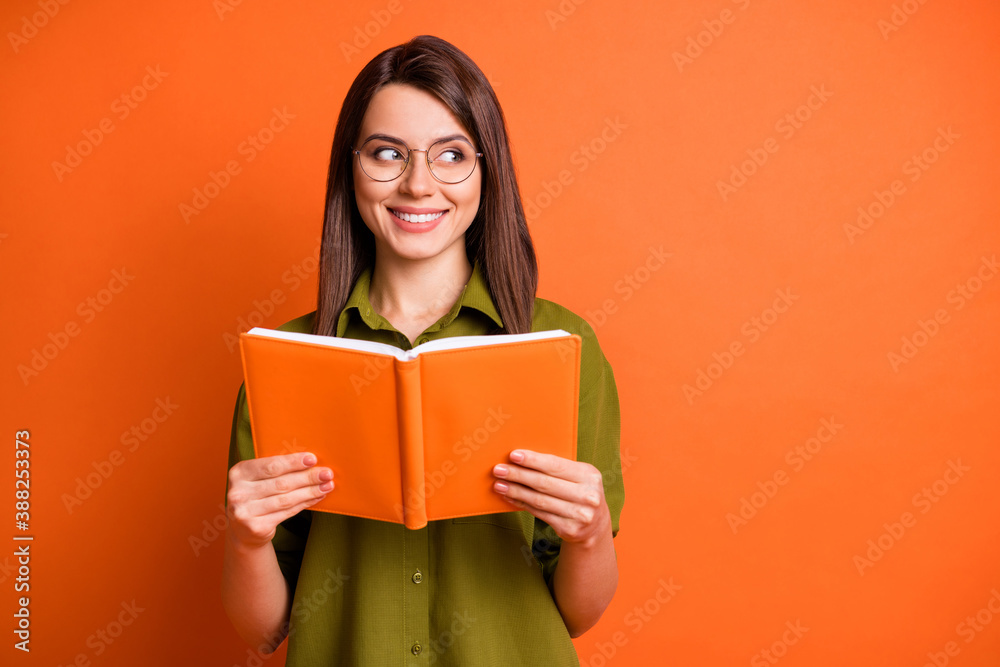 Photo portrait of curious female student wearing glasses keeping book looking at blank space isolated on vivid orange color background Photo portrait of curious female student wearing glasses keeping book looking at blank space isolated on vivid orange color background