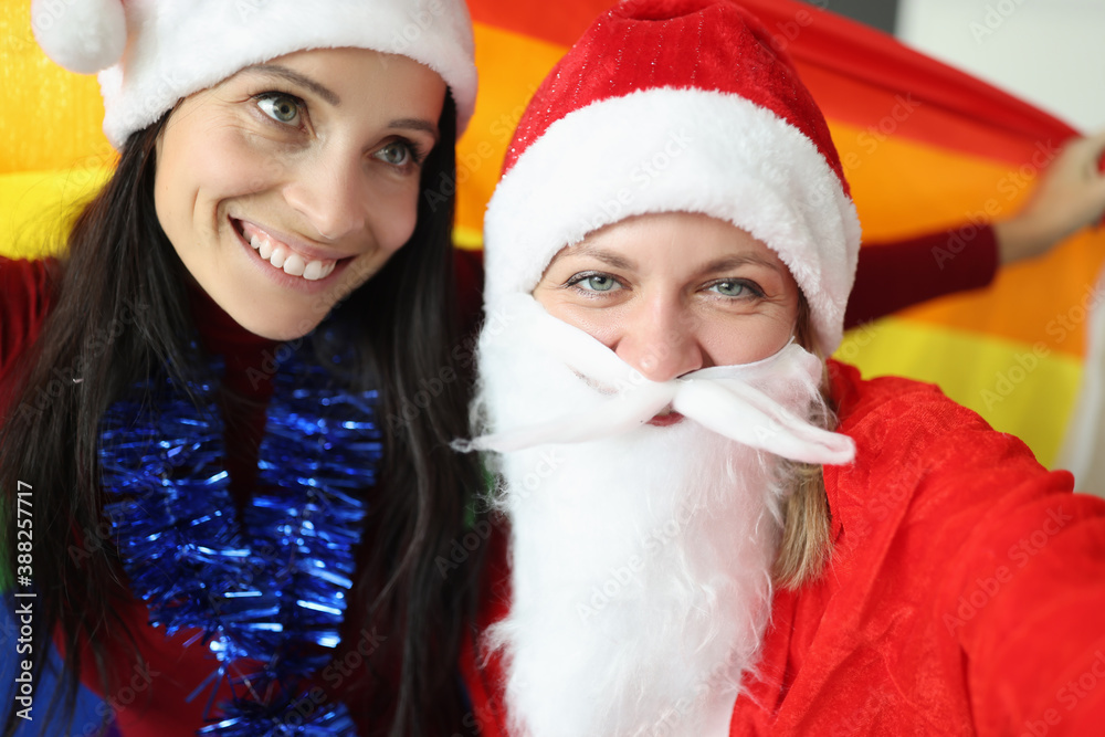 Fototapeta premium Two women in santa claus hats and santa claus costume hold lgbt flag. Christmas time and new year holidays and same sex marriage concept
