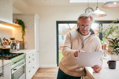 Принти на полотні Mature Man At Home In Kitchen Drinking Coffee And Looking At Financial Documents
