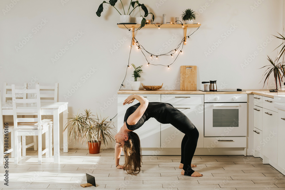 Woman of European appearance doing yoga in the kitchen. Sports at home online. A woman with long dark hair stands in a difficult yoga pose. - obrazy, fototapety, plakaty 