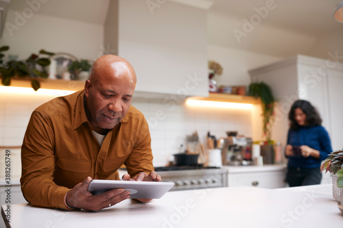 Mature Man At Home In Kitchen Looking At Digital Tablet With Female Partner In Background