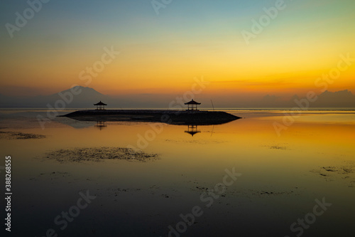 Sunrise seascape. Mountains and Agung volcano. Traditional gazebos on an artificial island in the ocean. Water reflection. Sanur beach, Bali, Indonesia.