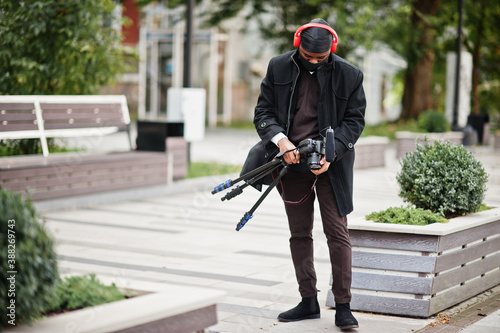 Young professional african american videographer holding professional camera with pro equipment. Afro cameraman wearing black duraq and face protect mask, making a videos. photo