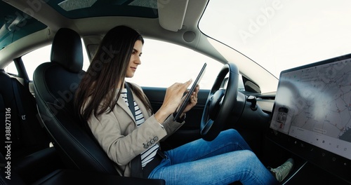 Side view of young Caucasian woman sitting in electric car and tapping on tablet. Joyful female scrolling and browsing on device in vehicle. Woman tapping on dashboard touch screen. Innovation concept