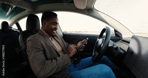 Concentrated young African American male sitting in modern electric car and browsing on computer. Handsome man holds smartphone and typing on laptop while traveling in autopilot vehicle. Tech concept