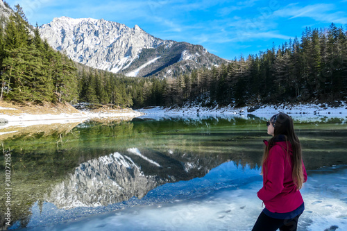 A woman walking around the shore of Green Lake, Austria. Powder snow covering the mountains and ground. Soft reflections of Alps in calm lake's water. Winter landscape of Austrian Alps. Calmness