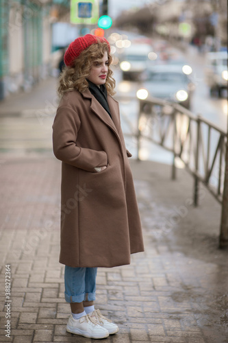 Cute girl in a red hat and brown coat is waiting near the road © Kiryakova Anna