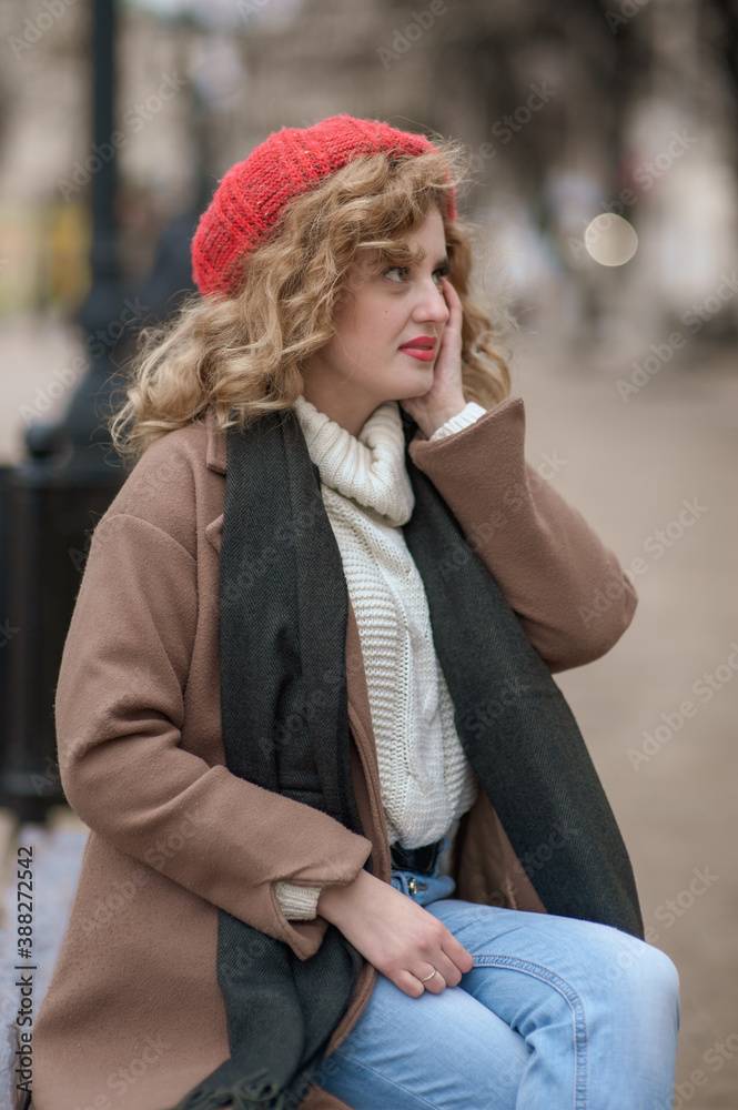 A cute girl in a red hat sits on a wooden bench on a city street A cute girl in a red hat sits on a wooden bench on a city street