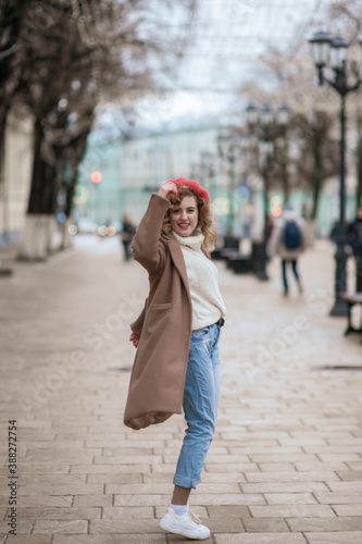 Beautiful brunette young woman in a red hat, jeans, and a coat happily walks along the street © Kiryakova Anna
