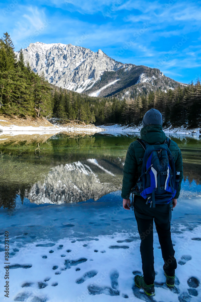 A man walking around the shore of Green Lake, Austria. Powder snow covering the mountains and ground. Soft reflections of Alps in calm lake's water. Winter landscape of Austrian Alps. Calmness