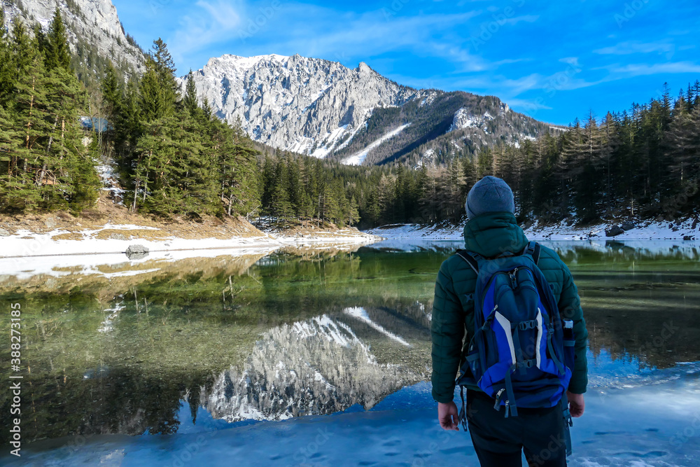 A man walking around the shore of Green Lake, Austria. Powder snow covering the mountains and ground. Soft reflections of Alps in calm lake's water. Winter landscape of Austrian Alps. Calmness