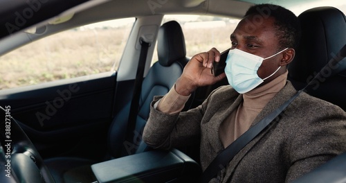 Young African American handsome man sitting in electric vehicle and speaking on cellphone. Close up of male in mask in ecological car chatting on smartphone. Quarantine concept © VAKSMANV