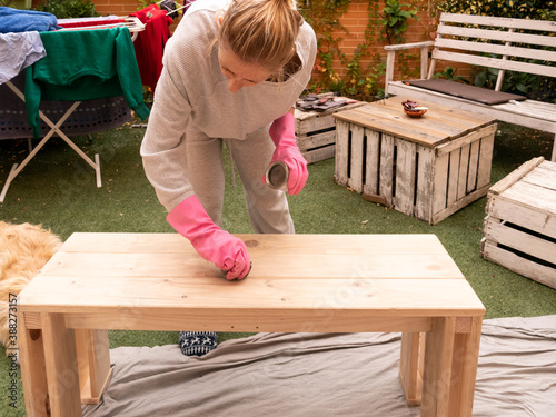 BLOND WOMAN IN PAJAMA, HOUSEWIFE, GIVING WAX TO WOODEN BENCH WITH PINK RUBBER GLOVES