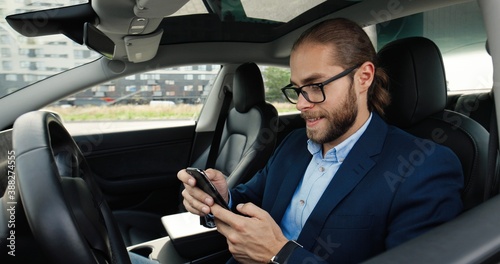 Close up of handsome Caucasian man driver in electric automobile smiling and searching internet on cellphone. Young happy male sitting in modern car and tapping on smartphone. City concept © VAKSMANV