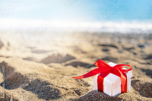 White gift box with a red ribbon standing on the sand with blue sea and sun on background. Holidays on the beach next to the sea.