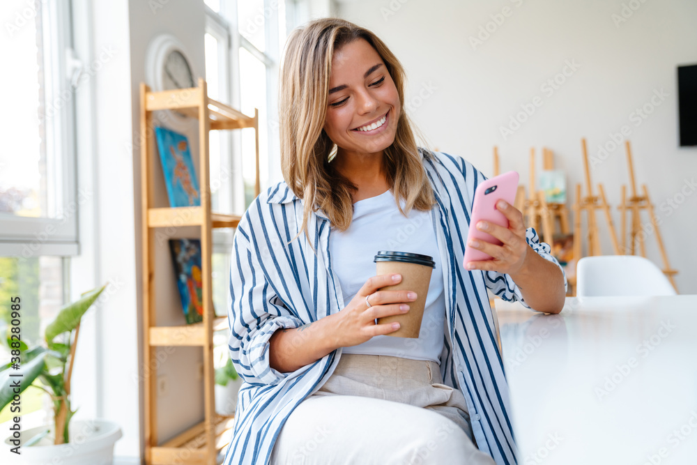 Young caucasian woman holding coffee cup and cellphone in bright room Young caucasian woman holding coffee cup and cellphone in bright room