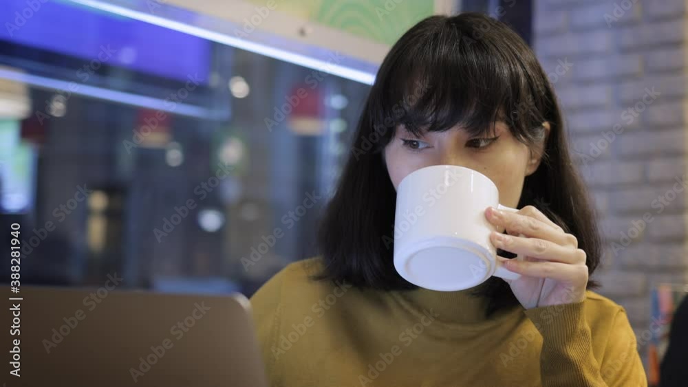 Young asian woman with laptop in cafe, girl drinking a cup of coffee working indoors. Gimbal shot closeup of student concentrated looking at the laptop in city cafe