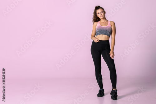 Young athletic woman standing in fitness outfit with ponytail in front of a pink background