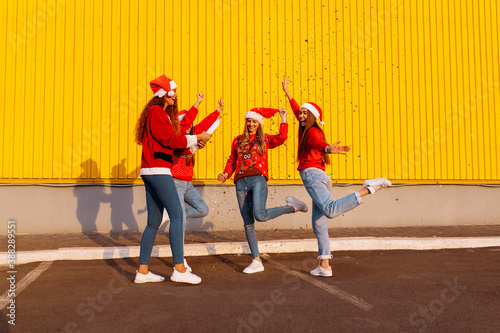 Happy young women wearing santa claus hats having fun and jumping with colorful confetti outdoors. New Year celebration
