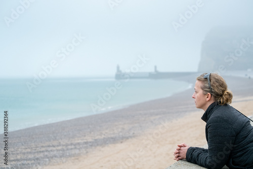 Woman wearing warm clothes and a purse looking out over a foggy sea from a pebble beach on the shore © Bjorn B