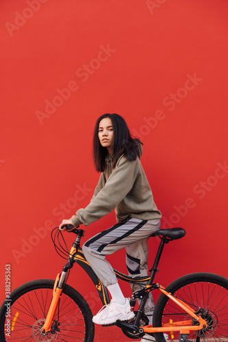 Fototapeta Naklejka Na Ścianę i Meble -  Portrait of young attractive woman on bicycle, isolated on red background, looking at camera with serious face. Copy space