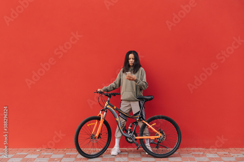 Fototapeta Naklejka Na Ścianę i Meble -  Pretty woman in sportswear stands with a bicycle on a background of a red wall and uses a smartphone with a serious face.