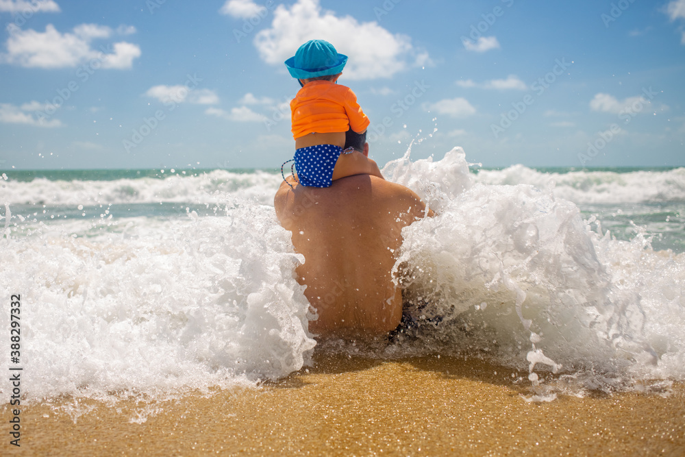 father's day. Dad protect. the baby sits on dad’s shoulders, dad sits on the beach and a wave comes.