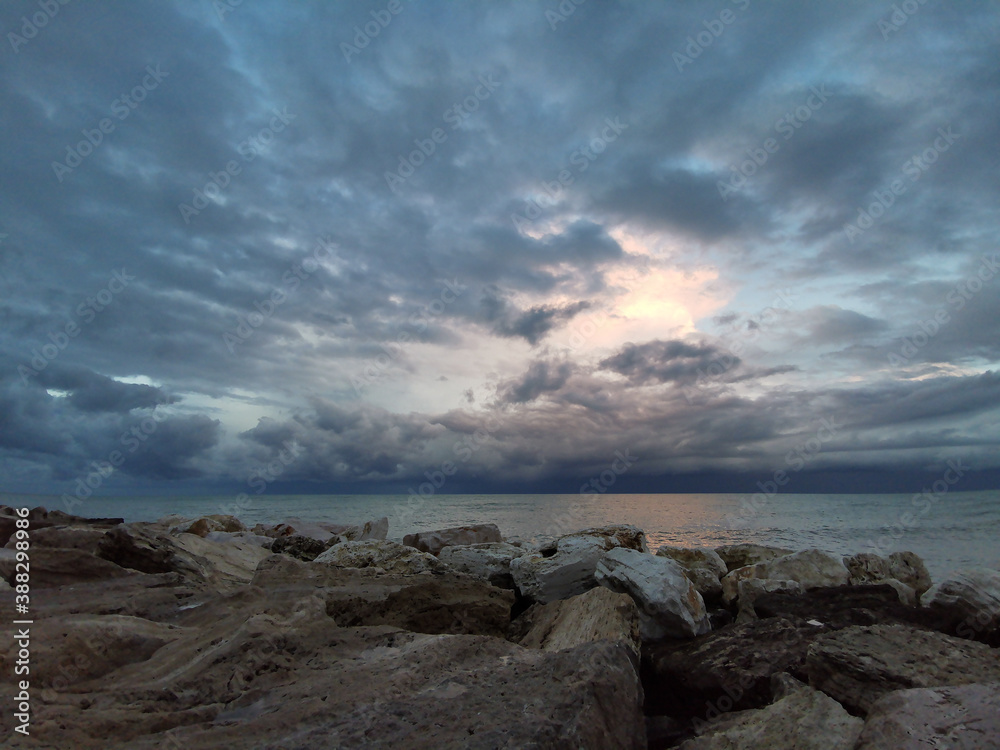 Tramonto sul mare Adriatico visto dagli scogli in una giornata invernale nuvole luminose e orizzonte scuro riflessi sull’acqua