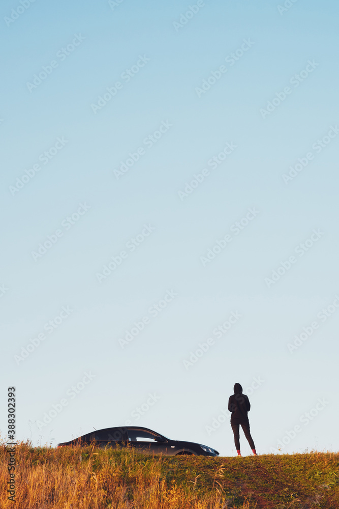 Fototapeta premium A girl in black clothes stands next to a black sedan car on a hill, against a dim sky, copy space. Concept of auto tourism and travel. Silhouette of a girl and a car at sunset in autumn