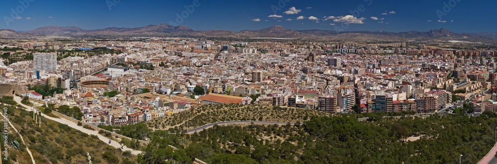 A viewpoint from a hill on the Spanish city Alicante and the mountains against blue sky. A panoramic view of a valley with green trees and roads in the forefront.