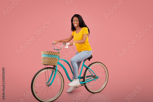 Fototapeta Naklejka Na Ścianę i Meble -  Full length portrait of beautiful African American woman riding vintage bicycle over pink studio background