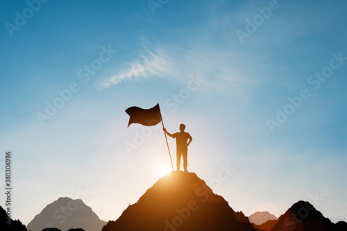 Silhouette of businessman holding flag on the top of mountain with over blue sky and sunlight. It is symbol of leadership successful achievement with goal and objective target. photo