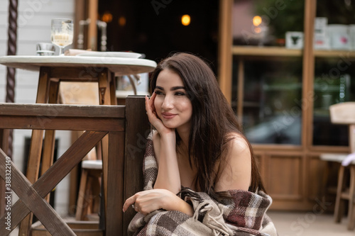 Beautiful girl resting sitting in cafe on the terrace, wrapped in a woolen plaid blanket. Autumn morning. Beautiful young woman with cup of coffee outdoors on autumn day