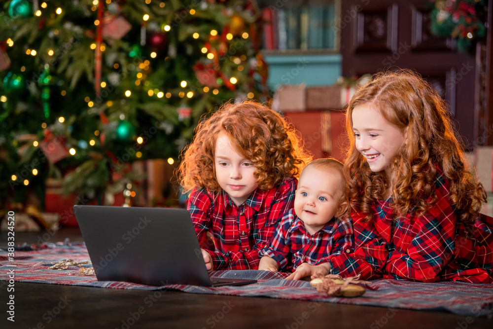 Funny red-haired children next to Christmas tree and gifts communicate online through a laptop. Funny red-haired children next to Christmas tree and gifts communicate online through a laptop.