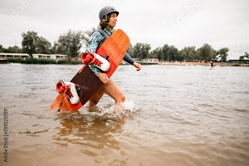 pretty woman with helmet and wakeboarding board walks on the water along the river bank photo