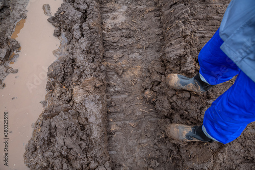 Feet of a man in a construction uniform. It stands on a muddy dirt road next to the footprint of the excavator tracks