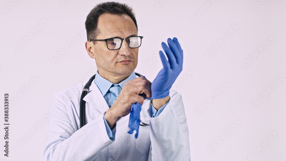 Serious handsome doctor in lab coat and stethoscope puts on latex protective gloves on isolated white background. Healthcare, medicine, health, medical specialist, clinic worker, protection, infection Serious handsome doctor in lab coat and stethoscope puts on latex protective gloves on isolated white background. Healthcare, medicine, health, medical specialist, clinic worker, protection, infection