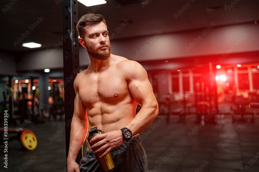 Portrait of athletic well built man with six pack abs. Half turned to the camera. Gym background. Guy holding bottle. Closeup. Portrait of athletic well built man with six pack abs. Half turned to the camera. Gym background. Guy holding bottle. Closeup.