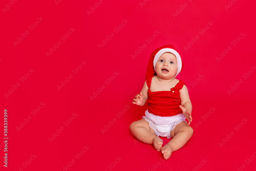 a small child of six months in a Santa hat sits on a red isolated photophone in expectation and hope, a place for text, the concept of the new year and holiday a small child of six months in a Santa hat sits on a red isolated photophone in expectation and hope, a place for text, the concept of the new year and holiday